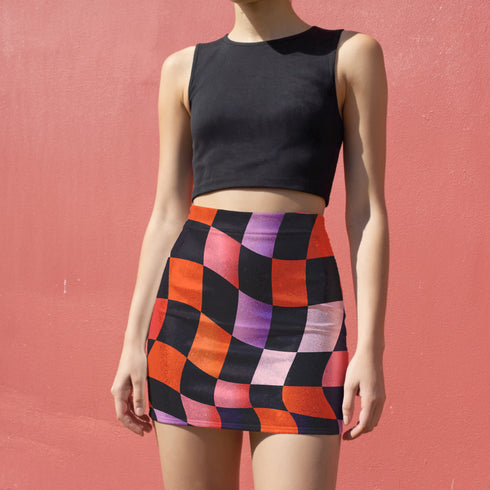 Honey Dazed mini pencil skirt featuring a bold wavy checkerboard pattern in black, orange, purple, and pink. Made from soft eco-polyester with a curve-hugging stretch fit and a flattering high-waist silhouette. The print has a slightly glossy finish, giving it a sleek and modern look. Designed for festival fits, streetwear moments, and retro rave energy. A vivid, sculptural mini skirt that pairs easily with crop tops and confident energy.